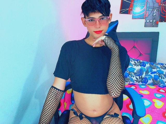 Allison, met kort haar en een bril, draagt een zwart topje, netruitjes en zwarte onderbroek, en zit op een kleurrijk bed, met haar hand op het kin, aantrekkelijk, zelfverzekerde pose, levendige kleuren, speelse sfeer.