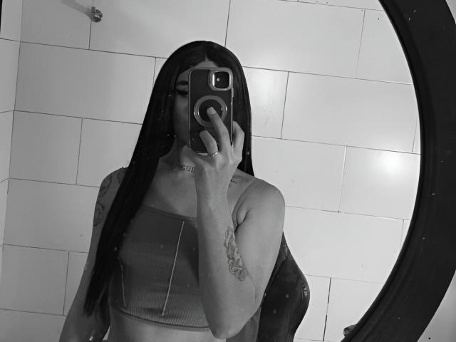 Selfie in bianco e nero di AdharaaMoon con lunghi capelli neri, che tiene un telefono davanti al viso, indossando un top corto a coste. persona con un tatuaggio ben visibile sulla parte superiore del braccio, in piedi di fronte a un muro piastrellato..