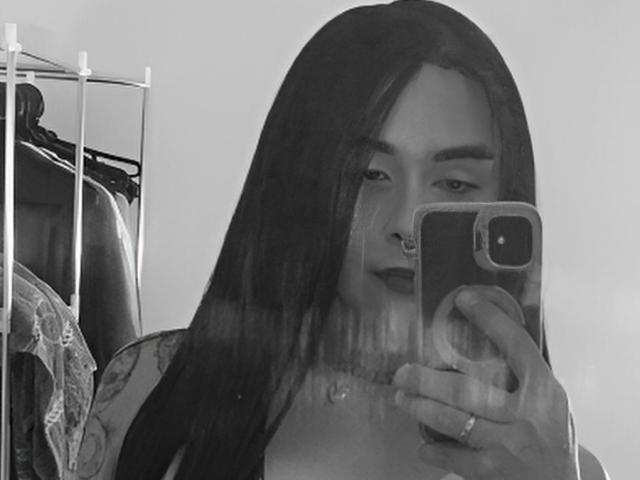 Schwarz-weißes Foto von AdharaaMoon, die ein Selfie vor einem Spiegel macht. Sie hat langes, dunkles Haar, dunklen Lippenstift und hält ein Telefon mit einer Ringhülle. Im Hintergrund sind Kleider an einem Kleiderständer zu sehen. Sexuelle Spannung entsteht durch ihren selbstbewussten Blick und den direkten Augenkontakt..