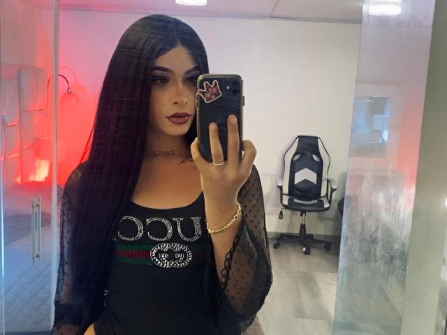 AdharaMoon, una donna attraente con lunghi capelli neri, si scatta un selfie allo specchio. Indossa una maglia nera trasparente con delle lettere scintillanti, rossetto scuro e una collana. Sfondo: sedia da ufficio, illuminazione rossa.