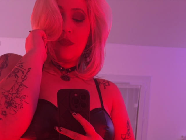 Fotografia di Blackwidows con capelli biondo platino, illuminazione rossa e tatuaggi sul braccio. Lei sta scattando un selfie, indossa un reggiseno nero, una collana e tiene in mano un telefono. Atmosfera rossa e seducente.