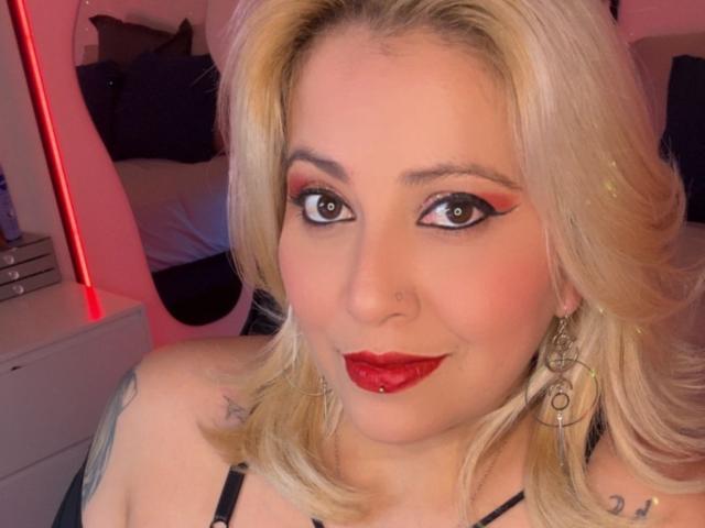 Close-up van Blackwidows, een blonde vrouw met rode lippenstift, opvallige oogmake-up en grote oorbellen. Ze heeft een tatoeage op haar schouder en draagt een zwarte top. Rood licht op de achtergrond.
