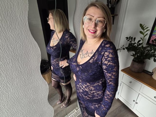 AmandalaFrancaise steht vor einem Spiegel und trägt ein schwarzes Spitzenkleid, feine Strumpfhosen und eine Brille. Das Spiegelbild zeigt ihr Profil. Sie lächelt, ihre Brüste sind deutlich sichtbar. Im Hintergrund befinden sich eine Topfpflanze und ein weißes Schrank..