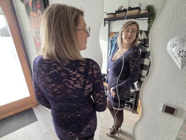 AmandalaFrancaise, con cabello rubio y gafas, vestida con un vestido negro de encaje, está de pie frente a un espejo, mirando. por sí misma. El reflejo en el espejo muestra su mirada segura. Es atractiva, con un escote visible..