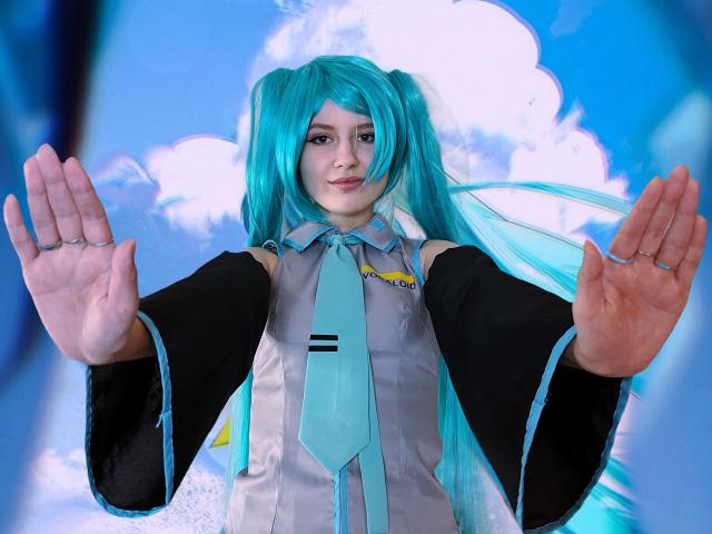 LinaVeritas, avec ses couettes turquoise, porte une tenue Vocaloid noire et argentée avec une cravate bleue, les mains levées, paumes vers l'avant. Avancer.
fond bleu et blanc vif, regard intense, léger sourire.