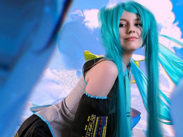 Fotografia di LinaVeritas, che fa cosplay di Hatsune Miku, con lunghi capelli turchesi, un abito nero e blu, un fiocco giallo e maniche staccate, sguardo rivolto verso l'alto con un sorriso appena accennato, sfondo di cielo blu..