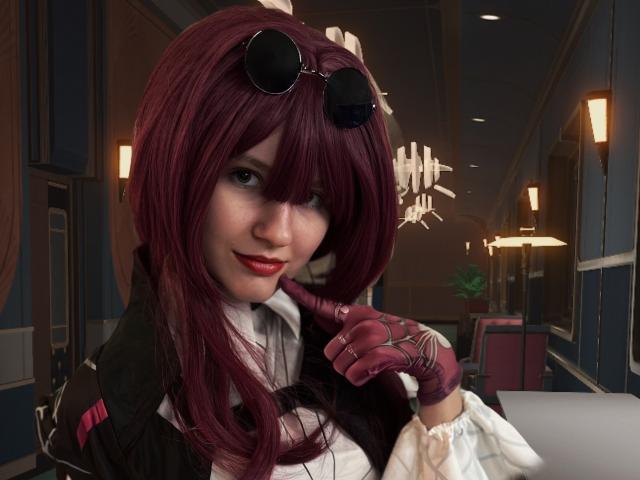 3Ein CGI-Bild von LinaVeritas, die als Cosplayer mit langen, dunkelroten Haaren, schwarzen Sonnenbrillen, einem weißen Hemd, einer schwarzen Weste und roten Handschuhen dargestellt wird. Nach vorne gebeugt, mit einem Grinsen, Restaurant im Hintergrund, schwach beleuchtet, warme Lichtleuchten..