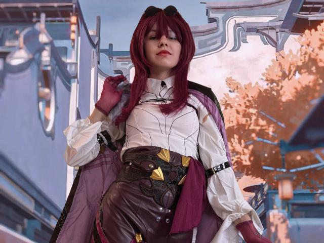 LinaVeritas se tient fièrement dans un décor futuriste, vêtue d'une chemise blanche ornée de motifs noirs et d'un pantalon marron. des détails dorés, et une cape violette. Elle a les cheveux roux et une expression sérieuse..