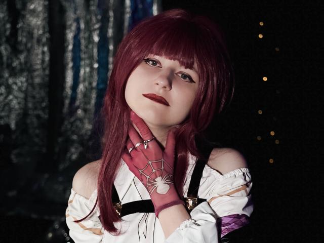 Photograph of LinaVeritas with deep red hair, bangs, and pale skin. She wears a white off-shoulder top, red gloves with spider web pattern, black strap, looking up with sultry expression.