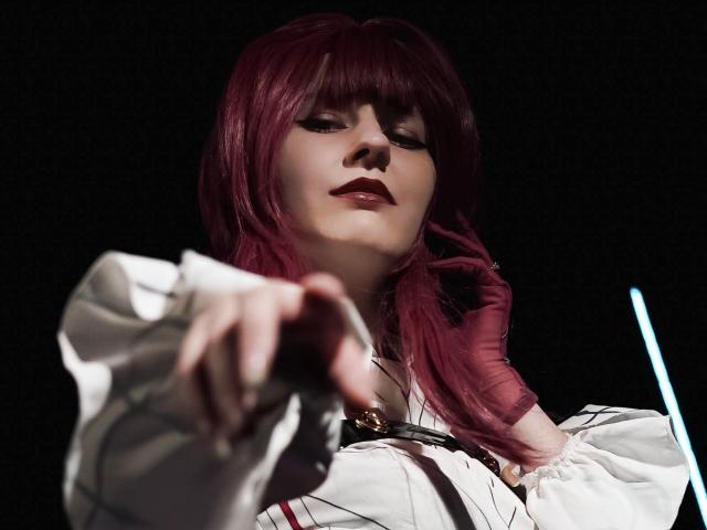 Photographie en gros plan de LinaVeritas, avec de longs cheveux roux, du rouge à lèvres foncé et une chemise blanche, pointant vers l'avant avec intensité. regard, éclairage tamisé, ombres dramatiques, tension sexuelle, premier plan flou, fond sombre.