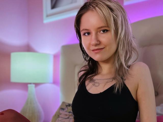 Fotografia de LinaVeritas com cabelo loiro na altura dos ombros, vestindo uma camiseta preta, sentada em um quarto com iluminação suave. Paredes em tons de rosa, uma luminária brilhante e iluminação roxa criam uma atmosfera sensual..
