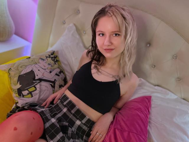 LinaVeritas, con i capelli biondo platino, indossa un top nero a tubino e una gonna a quadri, seduta su un letto con una testiera imbottita. È appoggiata all'indietro su cuscini rosa e gialli, e guarda verso la fotocamera..