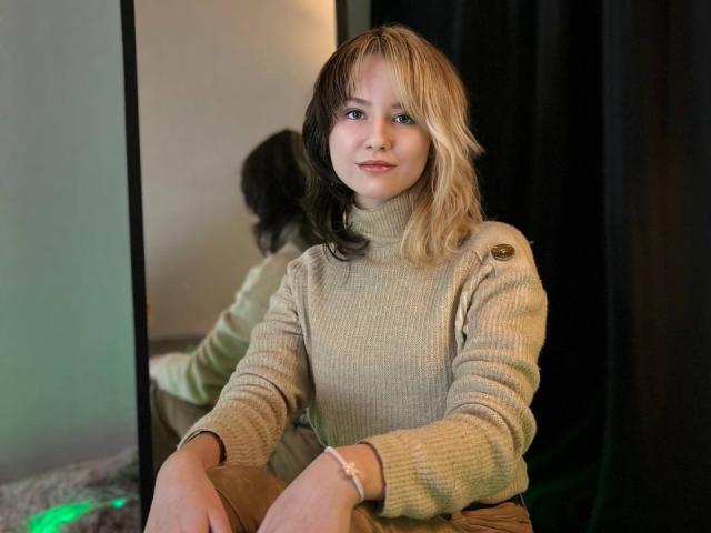 Fotografia di LinaVeritas, seduta con le gambe divaricate, che indossa un maglione beige a collo alto, con capelli biondi e neri, e un aspetto delicato. sorriso, sfondo scuro, specchio riflettente a sinistra, luce verde tenue.