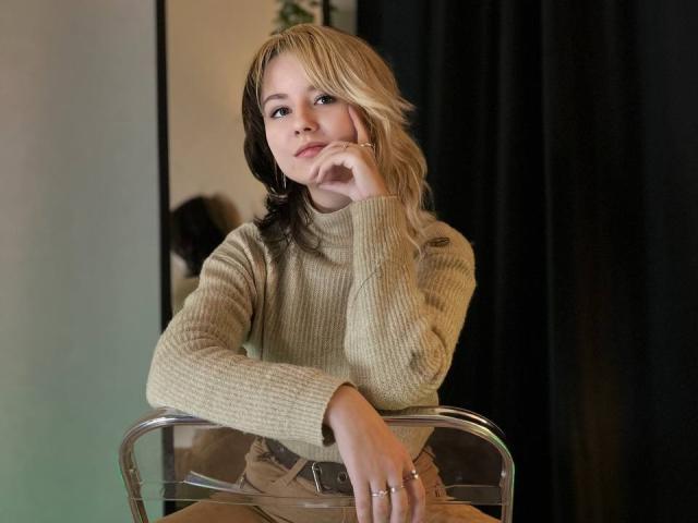 LinaVeritas, avec ses cheveux bruns clairs, portant un pull en tricot beige, est assise sur une chaise transparente, la main sur le menton, et regarde l'appareil photo. Lumière douce, fond noir avec un rideau. Ambiance sexy et pleine d'assurance..