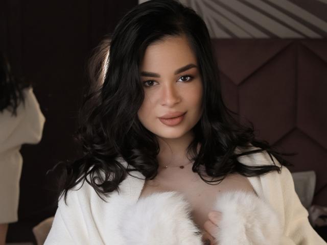 Photographie de LoraIvone avec de longs cheveux noirs, portant une robe en fourrure blanche, laissant entrevoir sa poitrine, avec des yeux sombres, des lèvres pulpeuses, et expression sensuelle, avec un arrière-plan faiblement éclairé. Tension sexuelle évidente..