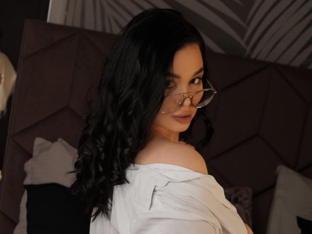 LoraIvone, avec ses longs cheveux noirs ondulés, portant des lunettes et une chemise blanche à une épaule, jetant un regard en arrière de manière séduisante. expression, fond sombre avec un motif géométrique, éclairage doux, tension sexuelle, regard séduisant.