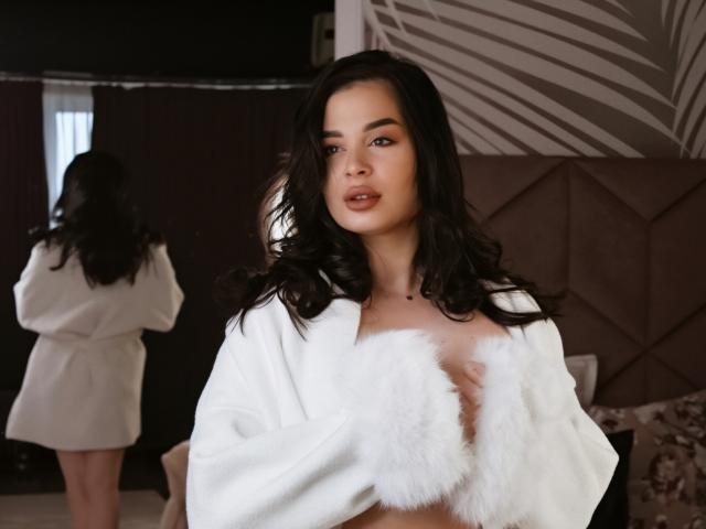 LoraIvone, une femme séduisante avec de longs cheveux noirs, porte un peignoir en fourrure blanche, laissant entrevoir une importante partie de sa poitrine. Réflexion dans le fond. Ambiance sombre et intime. Expression sensuelle et confiante. Extrêmement sexy.