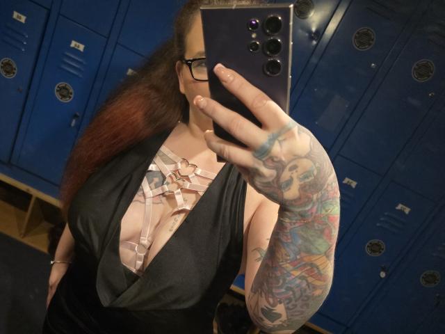 Selfie di JoyDiamond con lunghi capelli castani, vestito nero e scollatura accentuata con dettagli in pizzo floreale. Il braccio destro è ricoperto da numerosi tatuaggi e tiene in mano un telefono nero in uno spogliatoio blu. Assolutamente attraente..
