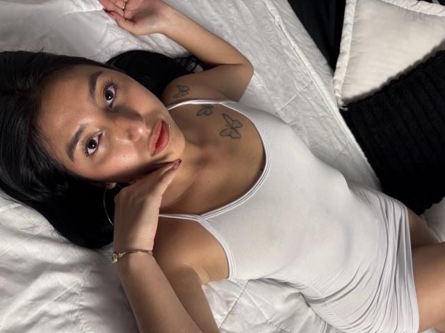 MiaLaurents lying on white pillows, wearing a tight white tank top, exposing her small breasts with butterfly tattoos. She gazes seductively, one hand near her face. Sunlight highlights her dark hair.