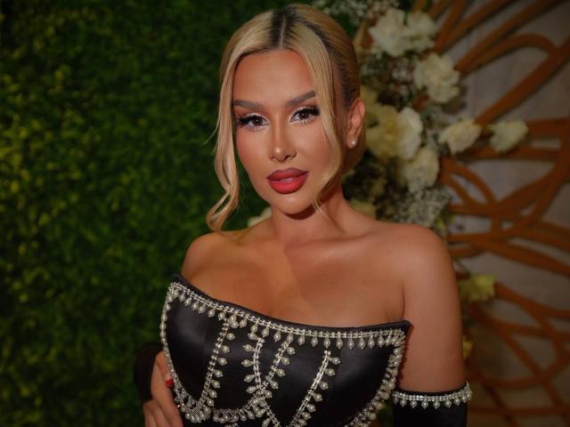 EvaZane, a sexy blonde woman with red lipstick, wears an off-shoulder black dress adorned with silver studs. She has a confident pose, green foliage and white flowers in the background.