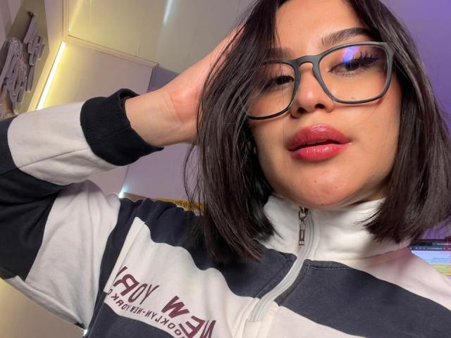 Nahaufnahme von AdaKolex, einer etwas molligen asiatischen Frau mit glattem, schwarzem Haar, die eine Brille mit schwarzem Rahmen und ein schwarz-weißes jacke mit Reißverschluss, Hand im Haar, rote, glänzende Lippen, anzügliche Mimik..