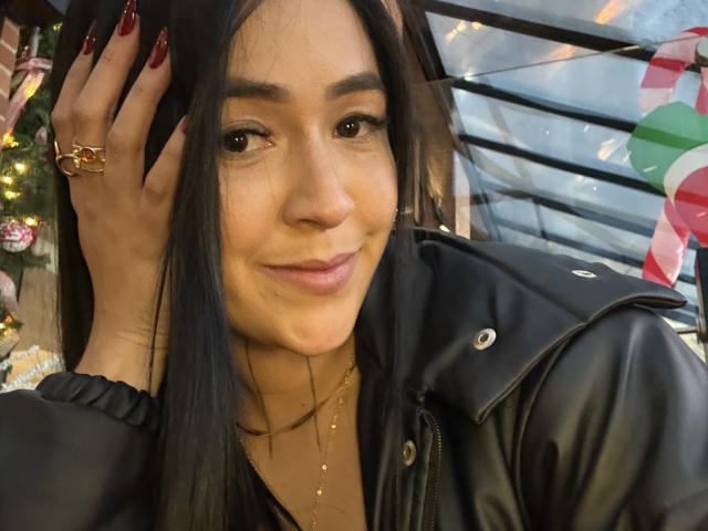 Close-up photo of NicoColeman, a young woman with long black hair, dark eyes, and a slight smile, wearing a black leather jacket, touching her hair with red nails, gold rings, and necklace. Fucking hot.