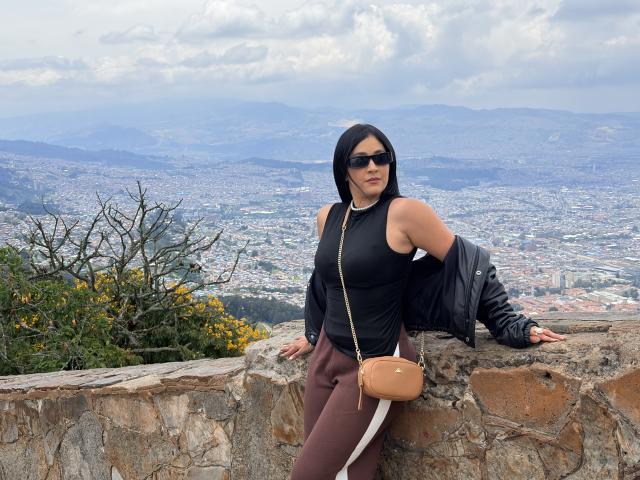 Nico Coleman, com cabelo escuro e óculos de sol, apoiado em uma parede de pedra, vestindo uma camiseta preta e calças marrons, com uma bolsa bege, com vista para uma extensa paisagem urbana. Tensão sexual evidente..