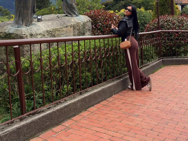 Nico Coleman se apoya en una barandilla de hierro forjado en un camino de ladrillo rojo, vistiendo una chaqueta de cuero negro y pantalones marrones. Con un bolso marrón y gafas de sol, mirando hacia arriba. Tensión sexual.