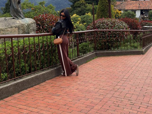 NicoColeman stands on a red brick patio, leaning against a black metal railing. She's wearing a black top, brown pants, and holding a yellow purse. Mountain and shrubs in background. Fucking sexy.