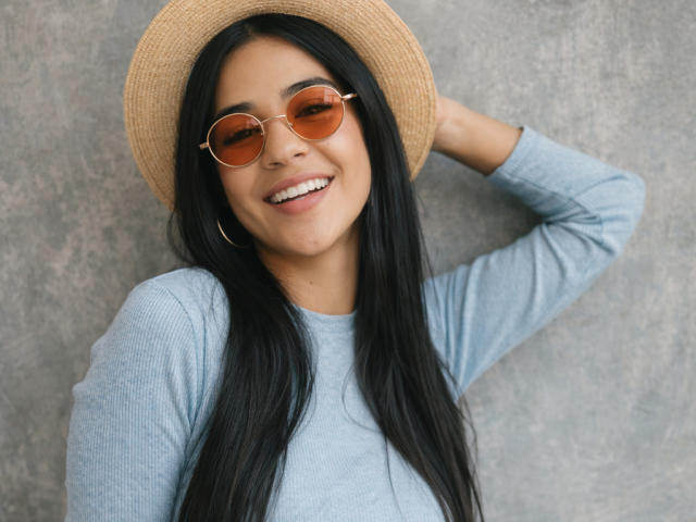 AmaiaCooper, a Latina woman with long black hair, wears a straw hat and round brown sunglasses, smiling with teeth showing. She's in a light blue sweater, hand behind her head. Fucking sexy.