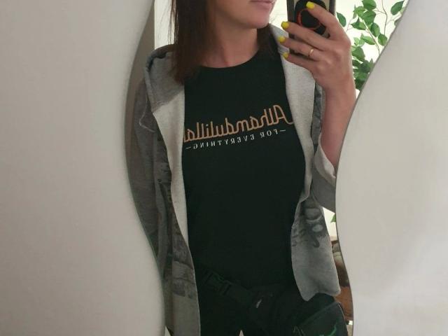 Fotografia de uma mulher com traços marcantes, segurando um telefone, vestindo uma camiseta preta com a inscrição "billionaire boyz", cinza. Jaqueta xadrez, calças pretas, unhas amarelas, encostada em uma parede branca..