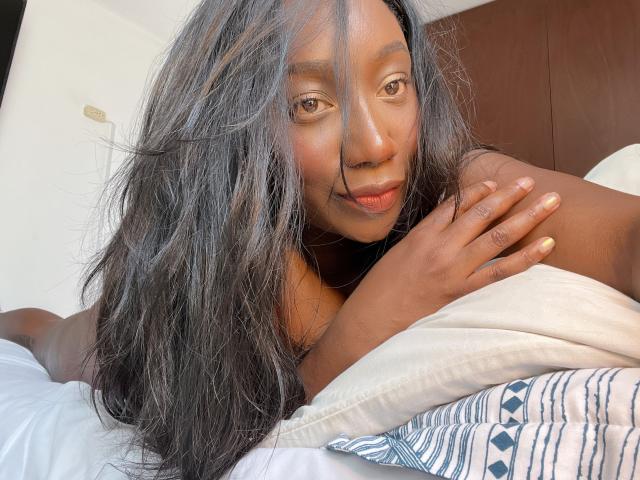 WarisEllen with dark brown skin, long black hair, and a subtle smile, lying on a white bed, her hand resting on her shoulder, her legs slightly spread, visible in bright light.