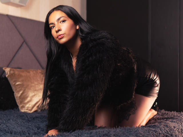 KiaraThomsomp on all fours on a blue shaggy bed, wearing a black fur coat and shiny black shorts. She has long black hair, sultry expression, and strong sexual tension. Sunlight highlights her.