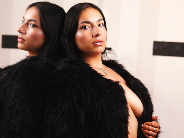Photograph of KiaraThomsomp, dual reflection, black fur coat, partially open, revealing small breasts, dark hair, serious expression, white background, high contrast, sexual tension, bold, confident, stylish, modern, seductive.