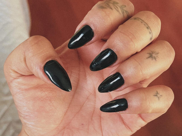 Primer plano de la mano de Elizabeth, con uñas negras brillantes y en forma de almendra. Su piel tiene tatuajes pequeños en los dedos. Los dedos están ligeramente curvados, mostrando las uñas de forma destacada. El fondo está desenfocado..