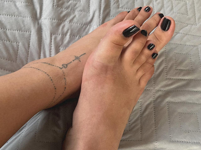 Primer plano de los pies de Elizabeth, con las uñas de los dedos de los pies pintadas de negro, tobillos cruzados y un tatuaje minimalista en su tobillo izquierdo. El tono de piel es claro, y el fondo es una colcha blanca acolchada..