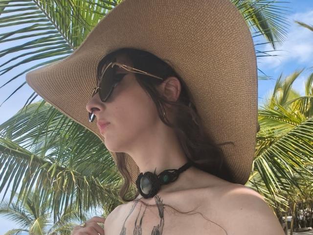Fotografía de KizzuKyuwu, una mujer de piel clara con cabello castaño oscuro, que lleva un sombrero de paja de ala ancha y de color oscuro. gafas de sol, sobre un fondo de vibrantes hojas de palmera verdes. Su pecho está desnudo, mostrando pequeños senos. Lleva un collar negro con un colgante grande, y sus ojos están medio cerrados. La luz del sol resalta sus rasgos..