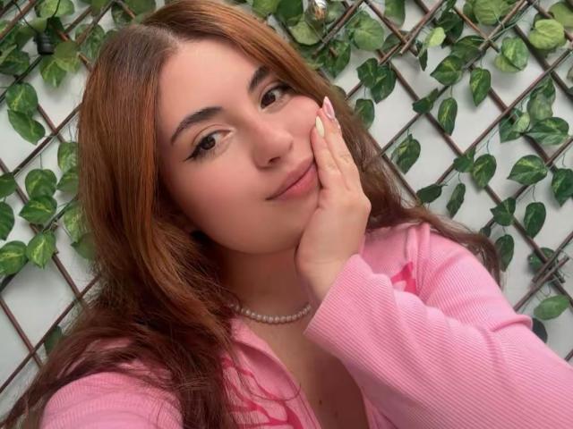 VicluX with long brown hair, light skin, and a slight smile, touching her cheek with one hand. Wearing a pink cardigan, pearl necklace, and large hoop earrings. Lush green leaves background.