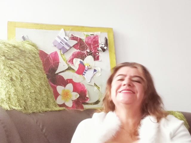 FuegoSexy has medium-length brown hair, smiling with eyes closed, wearing a white fluffy top, sitting on a brown sofa with a green pillow and colorful collage in background.