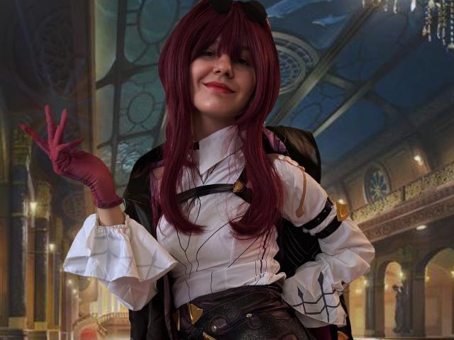 LinaVeritas steht selbstbewusst da und zeigt mit ihrer rechten Hand ein Friedenszeichen. Sie hat langes, dunkelrotes Haar, rote Handschuhe und ein schwarz-weißes Cosplay-Kostüm. Schwach beleuchteter, aufwendig gestalteter Innenraum im Hintergrund.