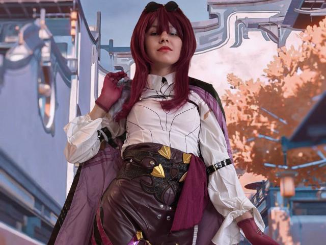 LinaVeritas se encuentra con confianza en un escenario de ciencia ficción, vistiendo una blusa blanca y detallada con mangas abullonadas, pantalones marrones y una capa color burdeos. Ella tiene el pelo rojo, y hay una tensión sexual evidente..