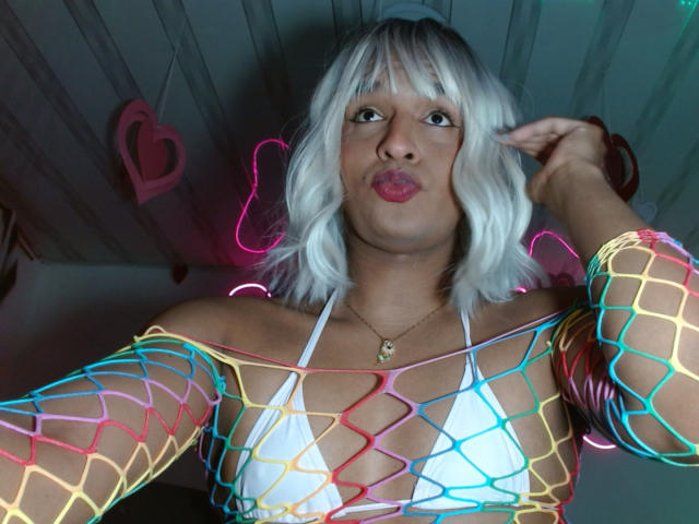 Photograph of IdiiaLatina with white bob haircut, wearing a neon fishnet top over a white bikini, glowing neon lights outline her body, pouty red lips, dark background with heart light.