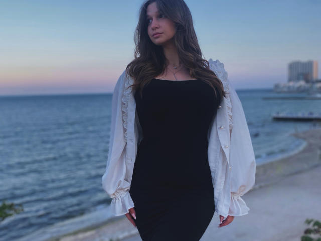 Miss Kuromi se tient sur une plage au crépuscule, vêtue d'une robe noire et d'une veste blanche. Elle a de longs cheveux bruns, un léger sourire, et on peut voir la forme de ses grandes seins. Fond avec vue sur l'océan et la ville..