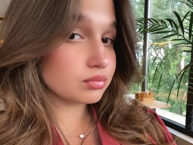 Close-up of MissKuromi with light brown, wavy hair, glossy pink lips, and a red shirt. She's wearing a delicate necklace. Background: bright window with green plants outside. Fucking sexy.