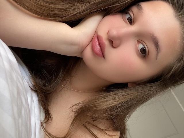 Close-up van MissKuromi, een jonge vrouw met een lichte huid en lang bruin haar, die haar hand op haar wang heeft. Ze heeft bruine ogen en roze lippen, en draagt een witte top. Ongelooflijk aantrekkelijk..