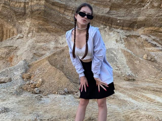 SenaCushingBerry stands against rocky terrain, wearing a white shirt, black skirt, and pearl necklace. She has braided hair, black sunglasses, and a white lace top. Leans forward, hands on thighs.
