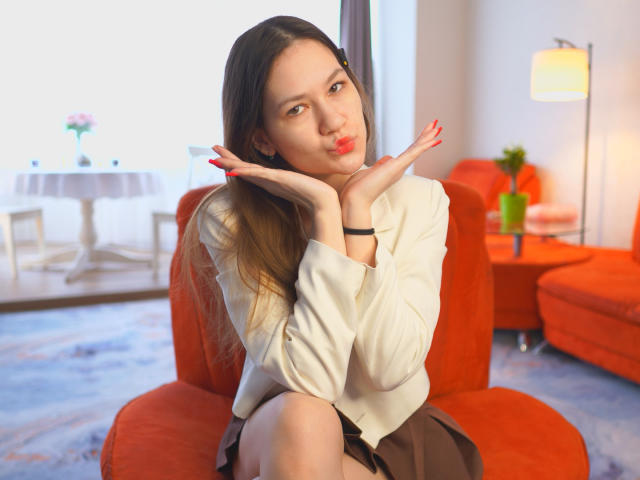 SenaCushingBerry sits on a bright red couch, hands framing her face, red lips slightly parted. Wearing a white blouse, brown skirt, long brown hair. Background: blurred modern living room.