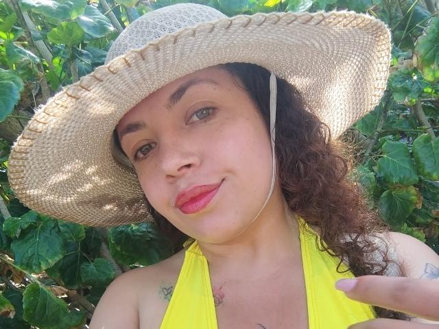 Foto ravvicinata di RubbyStrongX, una donna formosa con la pelle color nocciola e i capelli neri ricci, che indossa un cappello di paglia a tesa larga. Cappello e top giallo a fascia, in piedi in mezzo a un fitto fogliame verde. Con un sorriso appena accennato.