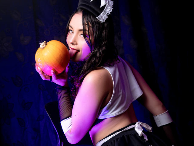 ViolettaMidnight hält eine leuchtend orangefarbene Kugel in der Hand, wobei ihr dunkles Haar und ihr schwarzes Outfit im Kontrast zu dem hellen Lila und Blau stehen. Beleuchtung. Sie leckt verführerisch an der Frucht, trägt dabei Netzstrümpfe und ein weißes Crop-Top..