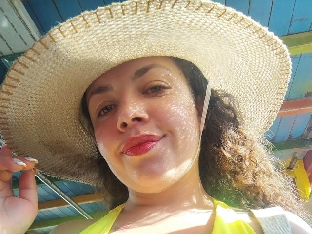 Close-up photo of RubbyStrongX with light brown skin, curly dark hair, and red lipstick, wearing a large straw hat, yellow top, under bright sunlight. Shadow on cheek, smiling subtly.